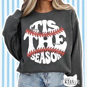May include: Dark gray sweatshirt with the text "TIS THE SEASON" in a baseball design. The baseball graphic is white with red stitching. The sweatshirt has long sleeves and a crew neck.