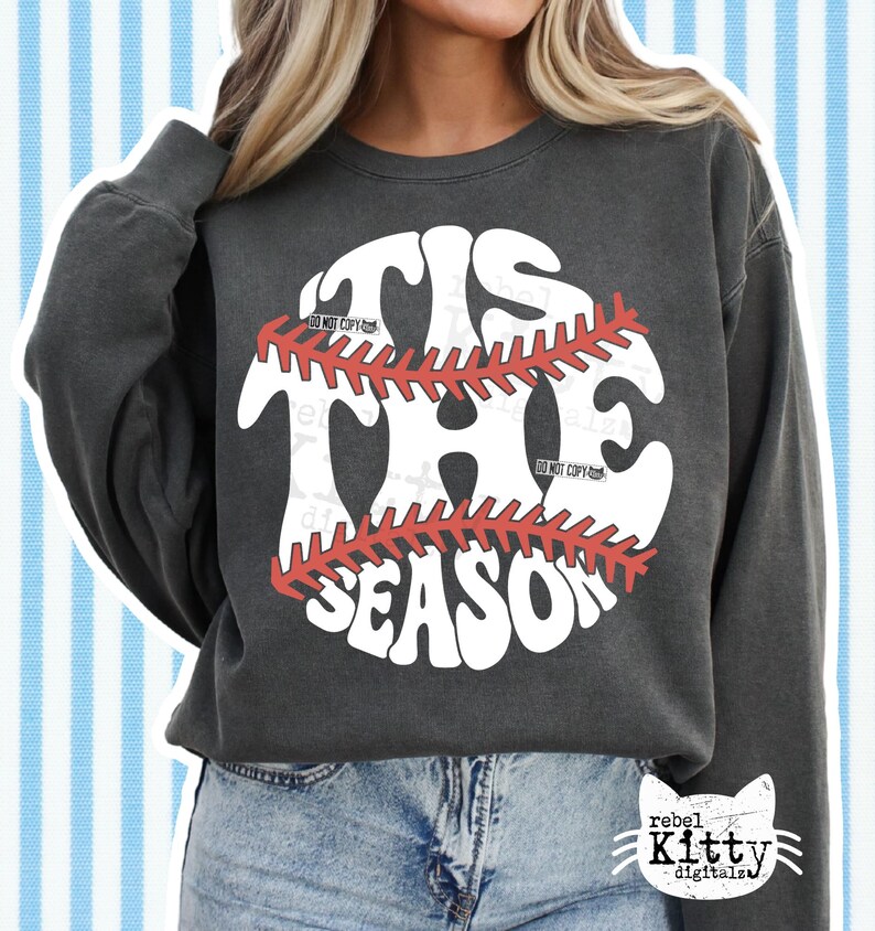 May include: Dark gray sweatshirt with the text "TIS THE SEASON" in a baseball design. The baseball graphic is white with red stitching. The sweatshirt has long sleeves and a crew neck.
