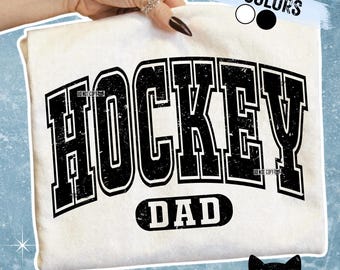 Puede incluir: Camiseta de color crema con las palabras "HOCKEY DAD" en una fuente negra desgastada. La camiseta también presenta el texto "IN COLORS" con opciones de color y el texto "*DISTRESSED & NON DISTRESSED*". Un logotipo de gato negro está en la esquina inferior derecha.