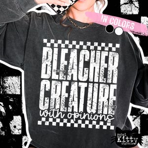 May include: Dark gray sweatshirt with the text "BLEACHER CREATURE with opinions" in distressed white lettering. The design includes a checkered pattern and a small black cat logo. The sweatshirt has a vintage, worn look.