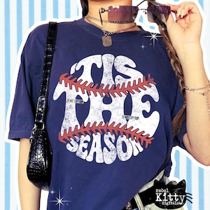 May include: Navy blue t-shirt with the text "TIS THE SEASON" in a distressed white font, overlaid with a baseball stitch design in red. The shirt has a vintage look.