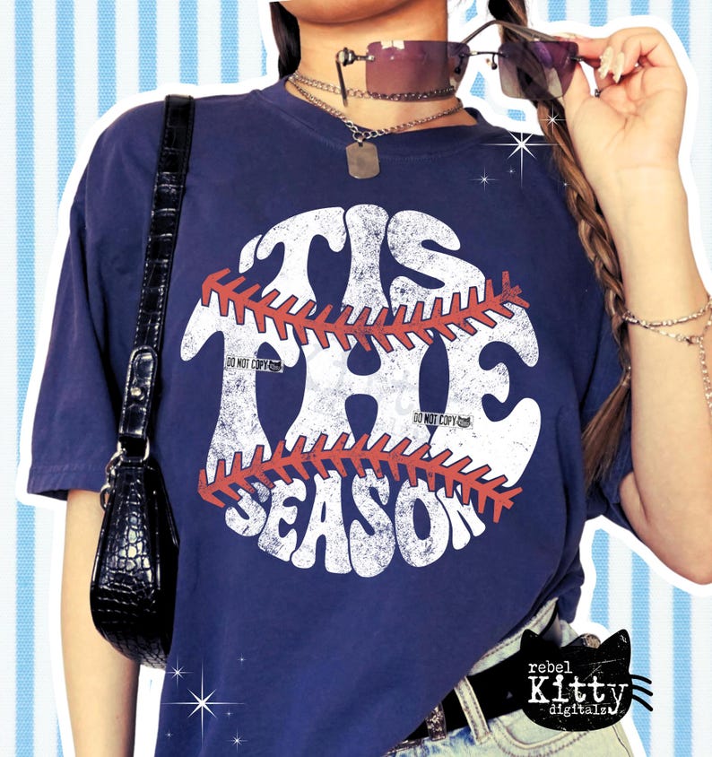 May include: Navy blue t-shirt with the text "TIS THE SEASON" in a distressed white font, overlaid with a baseball stitch design in red. The shirt has a vintage look.