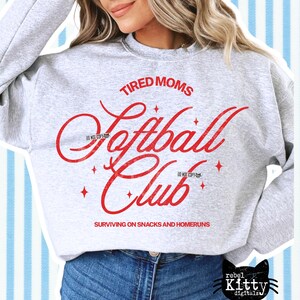 May include: A light gray sweatshirt with the words "TIRED MOMS Softball Club" in red script. The text "SURVIVING ON SNACKS AND HOMERUNS" is below the club name. The sweatshirt has a crew neck and long sleeves.