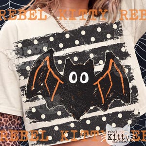 May include: A cream-colored t-shirt featuring a graphic of a black bat with orange wing outlines. The bat is centered over a black rectangular background with white polka dots. The text REBEL KITTY is printed in orange across the top and bottom.