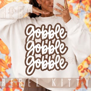 May include: A white long-sleeve sweatshirt featuring the text Gobble Gobble Gobble in a stacked, brown, distressed-style font. The lettering is bold and rounded, centered on the chest of the garment, creating a clear and legible graphic design.