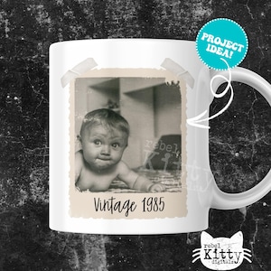 May include: White ceramic mug featuring a vintage black and white photo of a baby with the text "Vintage 1985". The mug has a handle and a teal "PROJECT IDEA!" sticker. The mug is a product of rebel Kitty digitals.