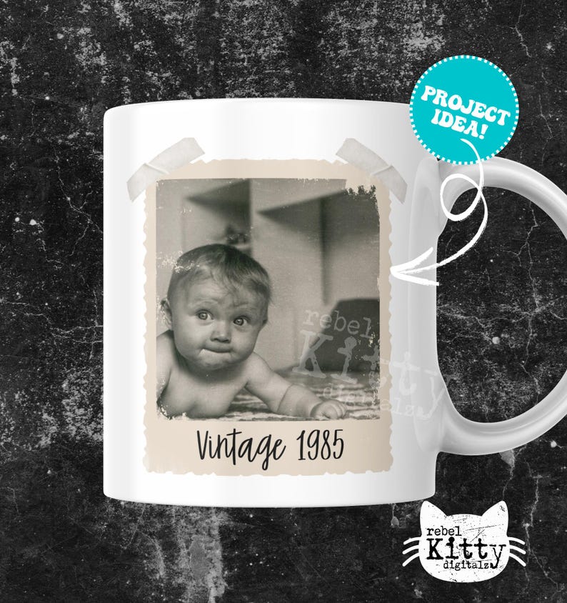 May include: White ceramic mug featuring a vintage black and white photo of a baby with the text "Vintage 1985". The mug has a handle and a teal "PROJECT IDEA!" sticker. The mug is a product of rebel Kitty digitals.