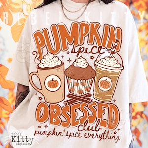 May include: An off-white t-shirt featuring a graphic of a coffee mug, a muffin, and an iced coffee cup with the text PUMPKIN SPICE OBSESSED CLUB and pumpkin spice everything. The design includes orange and brown colors with small star accents.