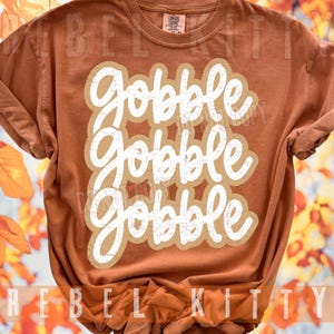 May include: A rust-colored short-sleeve t-shirt featuring the white text "gobble gobble gobble" in a stacked, distressed script font. The shirt has a crew neckline and a label at the inner collar that reads "Comfort Colors".
