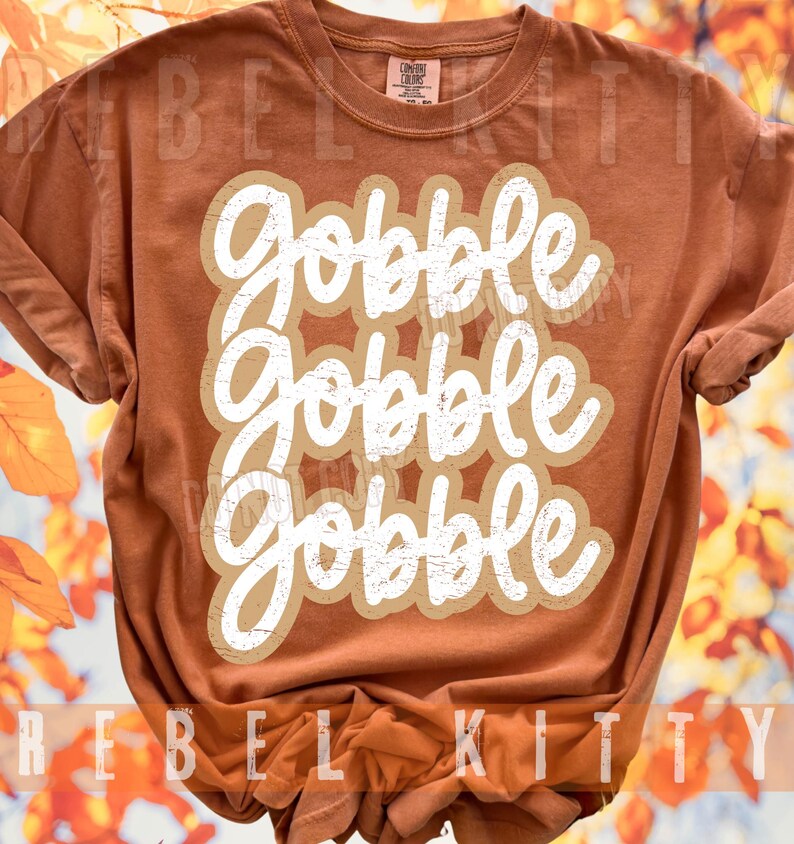 May include: A rust-colored short-sleeve t-shirt featuring the white text "gobble gobble gobble" in a stacked, distressed script font. The shirt has a crew neckline and a label at the inner collar that reads "Comfort Colors".