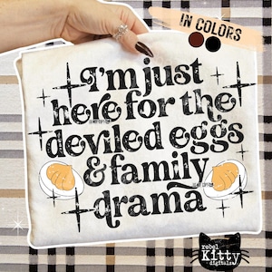 Könnte beinhalten: Cremefarbenes T-Shirt mit dem schwarzen Text "I'm just here for the deviled eggs & family drama". Das Design beinhaltet Illustrationen von gefüllten Eiern und Kreuzsymbolen. Das Markenlogo ist eine schwarze Katze.