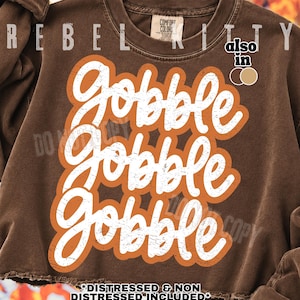 May include: A brown crewneck sweatshirt featuring the white text Gobble Gobble Gobble in a stacked, distressed script font. The text is outlined in a bold orange shape. The garment has a ribbed collar and a raw, unfinished hemline at the bottom.