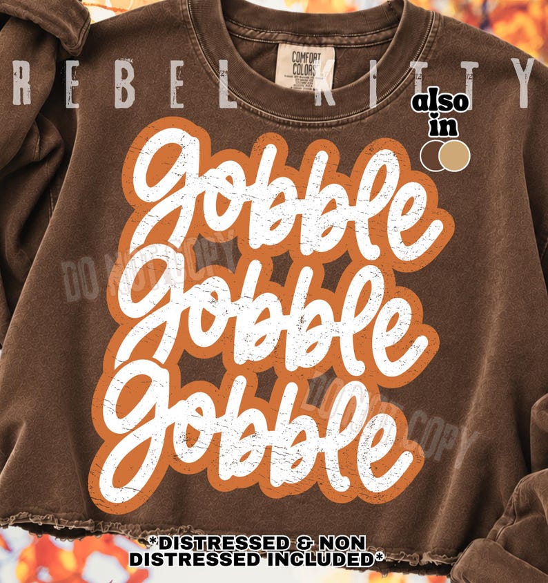 May include: A brown crewneck sweatshirt featuring the white text Gobble Gobble Gobble in a stacked, distressed script font. The text is outlined in a bold orange shape. The garment has a ribbed collar and a raw, unfinished hemline at the bottom.