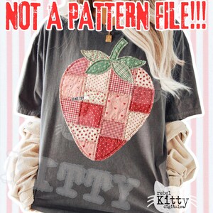 May include: Dark gray t-shirt featuring a patchwork strawberry design in shades of red and pink. The shirt has the text "NOT A PATTERN FILE!!!" in red at the top. The bottom of the shirt has the word "ITTY" in a faded font. The brand logo is in the bottom right corner.