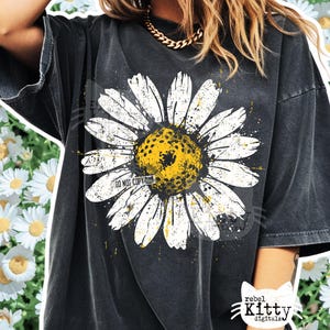 Puede incluir: Camiseta negra con un gran estampado de margarita. La margarita tiene pétalos blancos y un centro amarillo, con un efecto desgastado. El texto "DO NOT COPY" es visible en la margarita. La camiseta es de la colección Rebel Kitty Digitals.