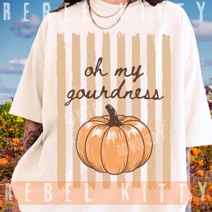 May include: An off-white short-sleeve t-shirt featuring a graphic print of a pumpkin centered over vertical tan stripes. The text "oh my gourdness" is printed in brown script above the pumpkin. The shirt has a crew neckline.