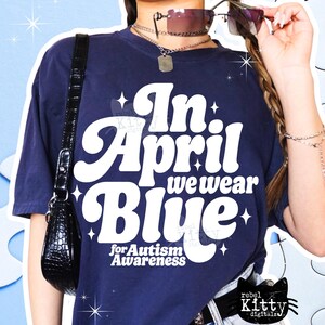 May include: Navy blue t-shirt with the text "In April we wear Blue for Autism Awareness" in white, retro-style lettering. The shirt has a relaxed fit and is adorned with star accents. A black purse hangs on the left side.