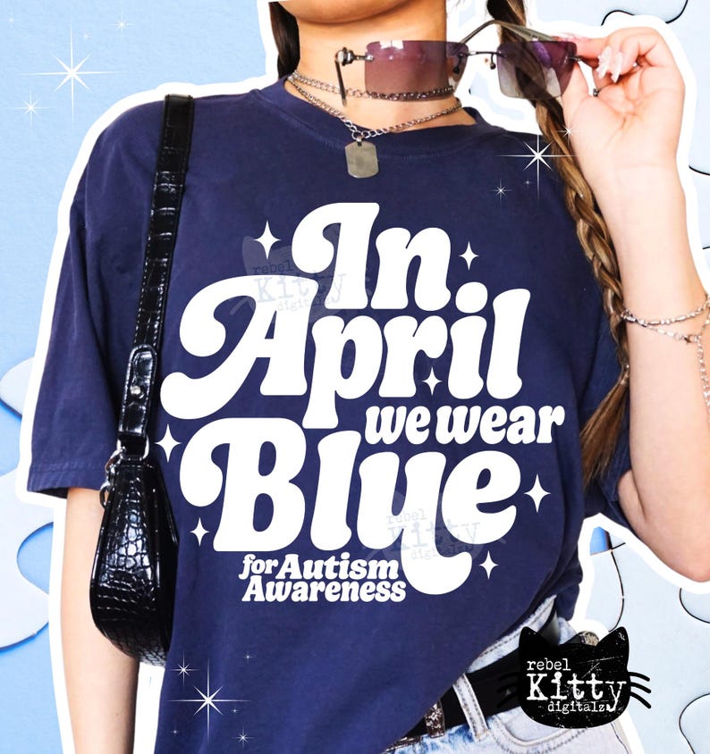 May include: Navy blue t-shirt with the text "In April we wear Blue for Autism Awareness" in white, retro-style lettering. The shirt has a relaxed fit and is adorned with star accents. A black purse hangs on the left side.