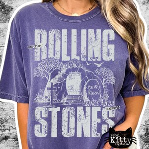 May include: Purple short-sleeved t-shirt with the words "ROLLING STONES" in large, distressed white letters. The design includes a cross, an open tomb, and the text "He is Risen! Since AD 33."