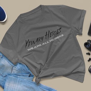 K&ouml;nnte beinhalten: Ein anthrazitfarbenes T-Shirt mit dem Aufdruck "XPINARY HEROES WE ARE ALL HEROES" in Schwarz und Wei&szlig;.