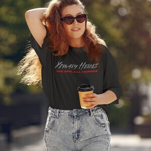 K&ouml;nnte beinhalten: Eine Frau tr&auml;gt ein schwarzes T-Shirt mit dem Text "Binary Heroes We Are All Heroes" in Rot und Wei&szlig;. Sie h&auml;lt eine braune Papptasse mit Kaffee.