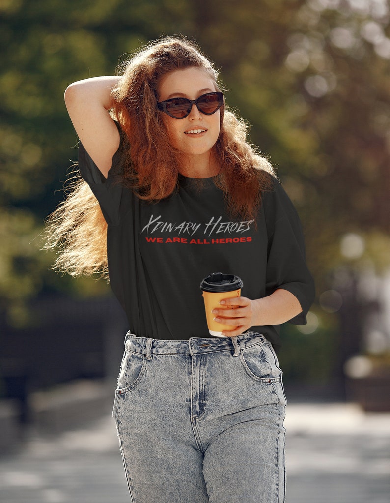 K&ouml;nnte beinhalten: Eine Frau tr&auml;gt ein schwarzes T-Shirt mit dem Text "Binary Heroes We Are All Heroes" in Rot und Wei&szlig;. Sie h&auml;lt eine braune Papptasse mit Kaffee.