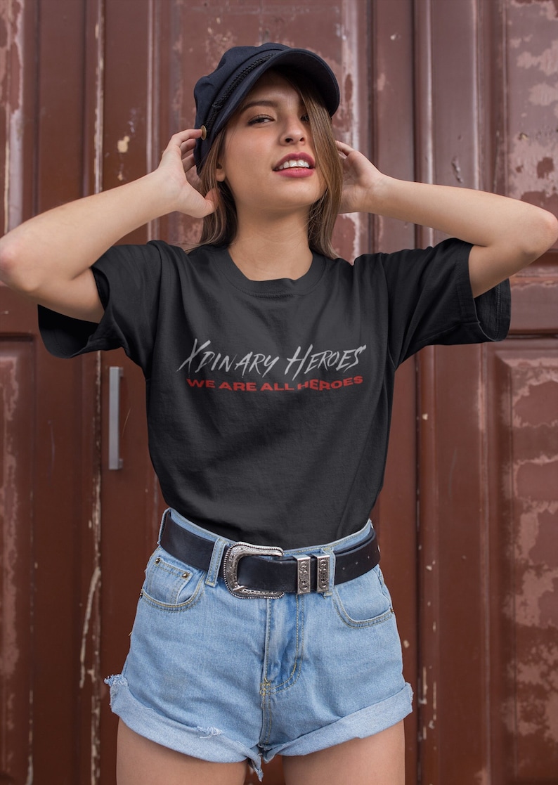 K&ouml;nnte beinhalten: Ein schwarzes T-Shirt mit wei&szlig;em Text, der "XPINARY HEROES WE ARE ALL HEROES" in einer stilisierten Schriftart lautet. Die Person, die das Shirt tr&auml;gt, tr&auml;gt auch eine schwarze M&uuml;tze und eine blaue Jeans-Shorts.