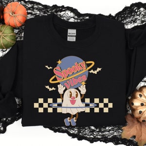 May include: Black sweatshirt featuring a graphic of a ghost holding a planet with the text "Spooky Vibes" on it. The ghost is wearing a red and pink headband and has a checkered pattern around it.