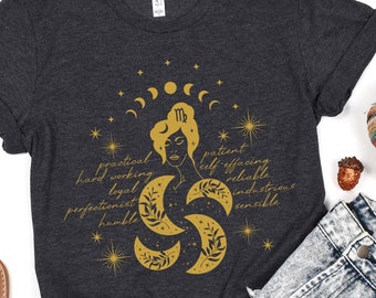 Puede incluir: Una camiseta gris oscuro con un gráfico dorado del signo del zodíaco Virgo rodeado de lunas y estrellas. El gráfico también incluye las palabras "práctico, trabajador, leal, perfeccionista, humilde, paciente, abnegado, confiable, industrioso, sensato".