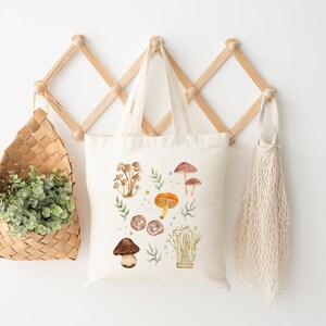 May include: A white canvas tote bag with a watercolor design of mushrooms and leaves. The bag is hanging on a wooden rack with a wicker basket and a string bag.