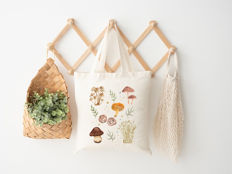 May include: A white canvas tote bag with a watercolor design of mushrooms and leaves. The bag is hanging on a wooden rack with a wicker basket and a string bag.