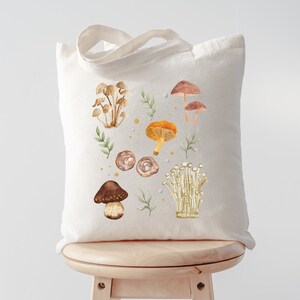 May include: A white canvas tote bag with a design of various mushrooms in shades of brown, orange, and white. The mushrooms are surrounded by green leaves and small yellow dots.