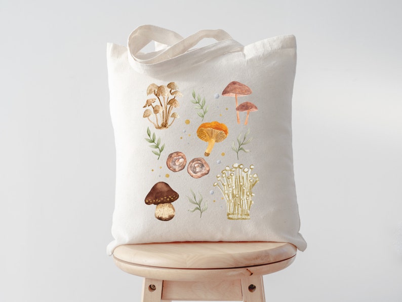May include: A white canvas tote bag with a design of various mushrooms in shades of brown, orange, and white. The mushrooms are surrounded by green leaves and small yellow dots.