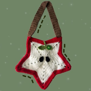 May include: A star-shaped crochet bag with a white centre and red edging. It has a brown strap and is adorned with green leaves and a small green bead. The bag's design is reminiscent of an apple.