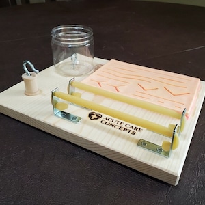 Puede incluir: Una tabla de entrenamiento de madera con una simulación de piel rosa y naranja, dos gomas elásticas amarillas, un gancho de metal y un frasco de vidrio transparente. La tabla está etiquetada con "ACUTE CARE CONCEPTS".