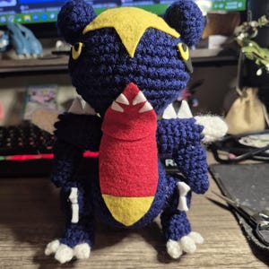 Può includere: Un peluche all'uncinetto fatto a mano a forma di creatura blu e gialla con una mascella inferiore rossa. Il giocattolo ha dettagli bianchi e si trova su una superficie di legno. La creatura ha una cresta gialla a forma di stella e piccoli occhi neri.