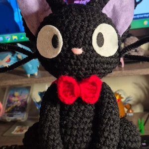 May include: A black crocheted cat plush toy featuring large white felt eyes, light purple inner ears, a small pink nose, and a bright red crocheted bow tie around its neck. The cat has long black whiskers extending from its cheeks.