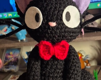 Puede incluir: Un peluche de gato tejido a ganchillo en color negro con grandes ojos de fieltro blanco, orejas de color violeta claro, una pequeña nariz rosa y una pajarita roja tejida a ganchillo alrededor del cuello. Tiene largos bigotes negros.