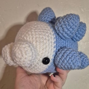 Puede incluir: Un peluche de dinosaurio hecho a mano con ganchillo, azul y blanco. El juguete tiene un cuerpo redondeado, una cabeza pequeña y una fila de púas azules en la espalda. Tiene ojos de seguridad negros. Hecho con hilo suave.