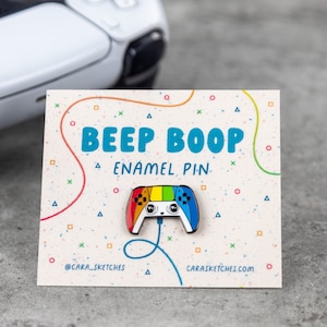 Puede incluir: Un pin de esmalte colorido con forma de mando de videojuego con un diseño de arcoíris y una linda cara de dibujos animados. El pin dice "Beep Boop Enamel Pin" e incluye el nombre de usuario de las redes sociales @CARA_SKETCHES y el sitio web CARASKETCHES.COM.