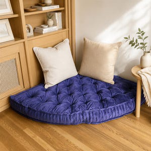 May include: A large, semi-circular, deep blue tufted floor cushion featuring a velvet-like texture and button detailing. Two square, beige-colored throw pillows are placed on top of the cushion, providing a contrast in color and fabric texture.