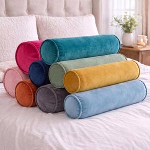 May include: A collection of nine cylindrical bolster pillows in various colors including teal, mustard yellow, light blue, sage green, navy blue, dark gray, burnt orange, magenta, and light pink, all featuring a soft, velvet-like fabric texture.