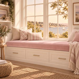 May include: A white window seat with three drawers featuring gold handles, topped with a long pink cushion. The seat is decorated with several pillows in solid cream, pink floral, and pink striped patterns, with a textured throw blanket nearby.