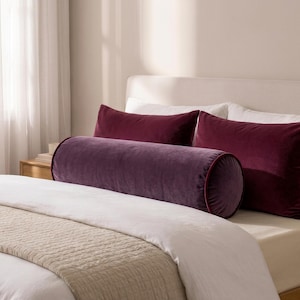 May include: A cylindrical bolster pillow in a deep purple velvet fabric with dark red piping, positioned in front of two rectangular deep purple velvet pillows on a bed with white bedding and a light beige textured throw blanket.