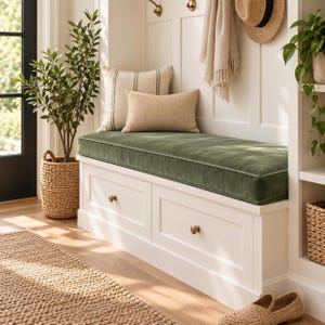 May include: A white wooden storage bench featuring two drawers with brass knobs and a long, rectangular olive green velvet cushion. The bench is topped with a square striped pillow and a rectangular beige pillow, positioned against a white wall.