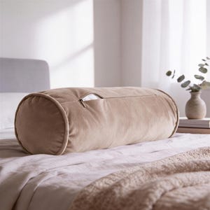 May include: A cylindrical bolster pillow in a solid light beige velvet fabric. The pillow features a horizontal zipper closure along the top side, revealing a small portion of white inner lining. The ends of the bolster are circular and piped.