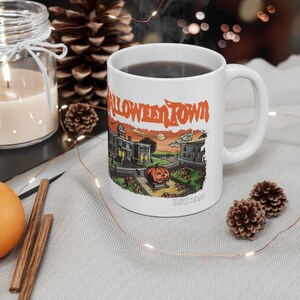 Puede incluir: Taza de cerámica blanca con una ilustración colorida de un pueblo espeluznante con una calabaza y el texto "Halloween Town Est. 1988".
