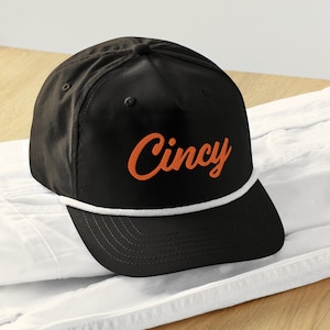 Puede incluir: Gorra de béisbol negra con ribete de cuerda blanca y la palabra "Cincy" bordada en naranja.