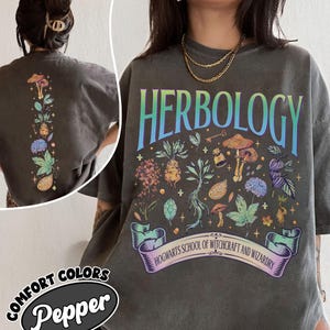 Peut inclure: Un t-shirt gris anthracite avec une impression graphique colorée portant le texte HERBOLOGY et HOGWARTS SCHOOL OF WITCHCRAFT AND WIZARDRY. Le motif comprend diverses illustrations botaniques, des champignons et de petites étoiles. Le dos est imprimé.