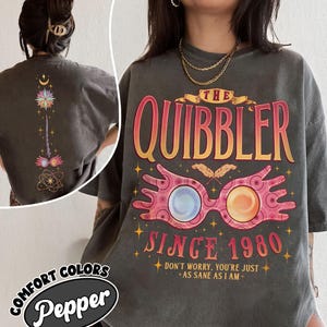 May include: A charcoal gray t-shirt featuring a graphic print of pink spectacles with blue and orange lenses. The text reads THE QUIBBLER SINCE 1980 and DON'T WORRY, YOU'RE JUST AS SANE AS I AM. The back shows a vertical design with a star and wand.